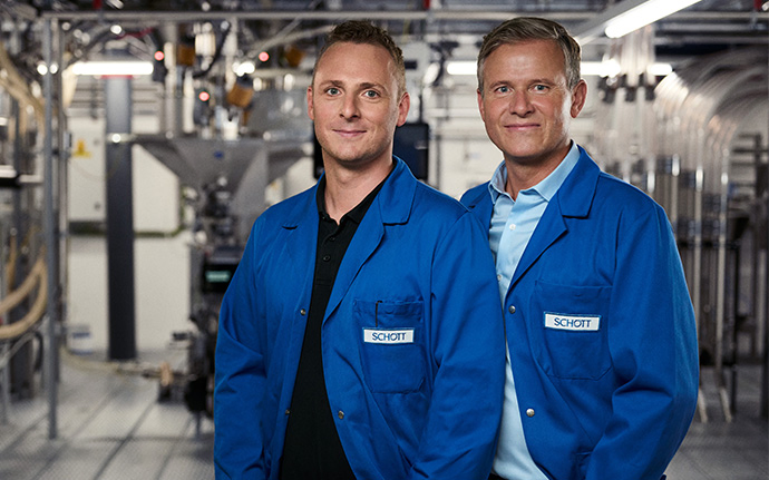 Zwei Personen in blauen Laborkitteln stehen in einer industriellen Produktionsanlage und blicken in die Kamera.