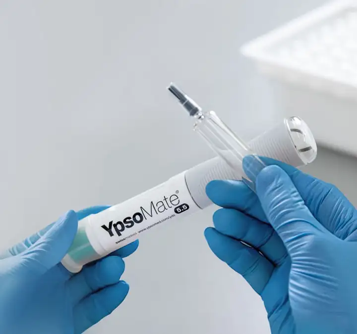 Gloved hands holding a YpsoMate® 5.5 autoinjector with syriQ BioPure® staked-needle glass syringe