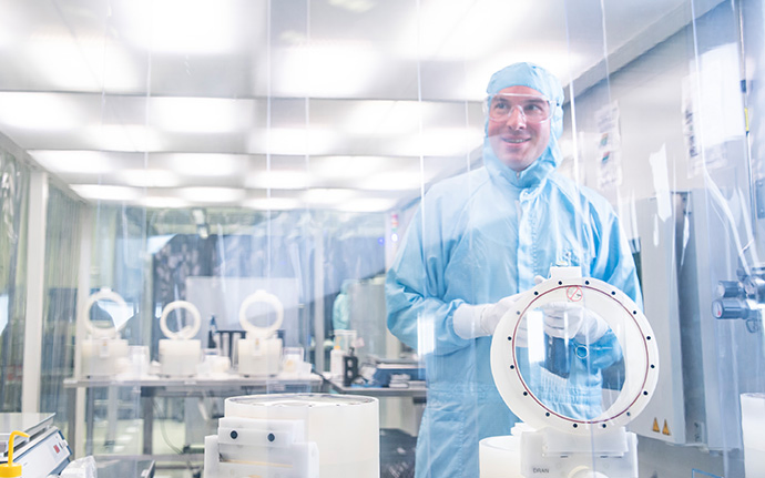 Eine Person in einem hellblauen Reinraumanzug arbeitet an einem runden technischen Bauteil in einem hochsterilen Laborumfeld.