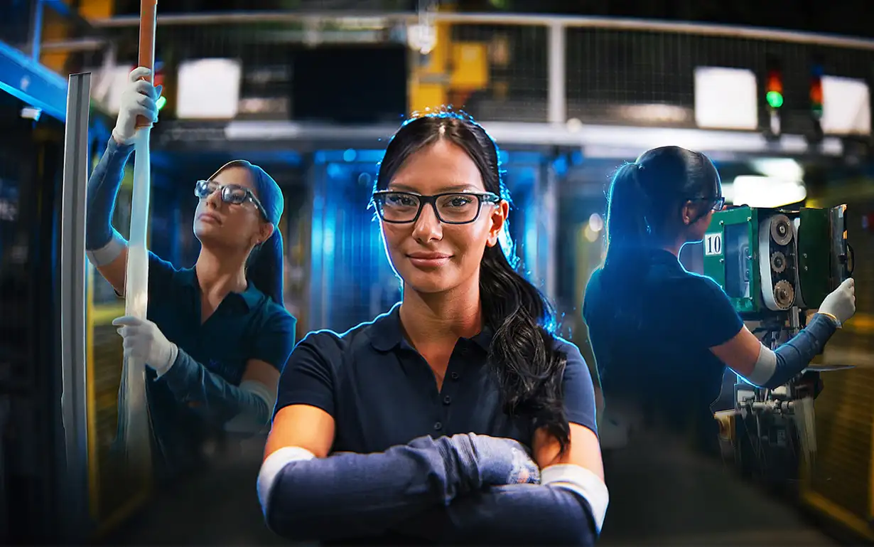 Frau in Arbeitskleidung und Schutzhandschuhen in einer Produktionshalle, umgeben von Maschinen und Metallstrukturen.