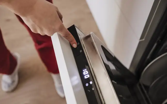 Person pressing the start button on a built‑in dishwasher with digital glass display.