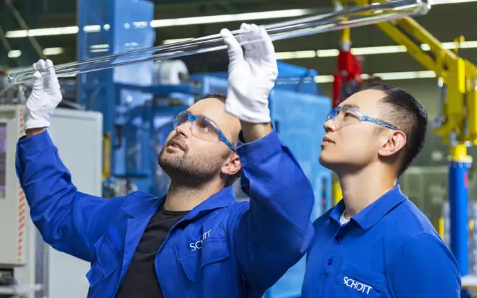 Zwei Männer in blauer SCHOTT Arbeitskleidung, einer hält ein Pharmarohr in den Händen, mit weißen Handschuhen.