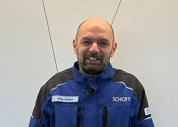Eine Person in blauer Arbeitsjacke mit Namensschild steht vor einer hellen Wand.