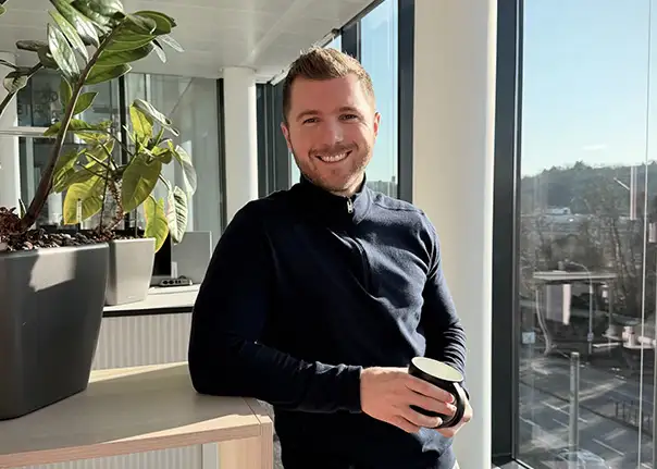 Eine Person lehnt mit einer Tasse in der Hand an einem Tisch in einem sonnendurchfluteten Bürobereich.