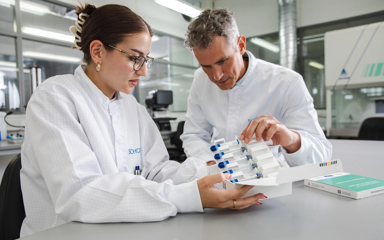 Woman and man in white coat with SCHOTT logo holding a cardboard box of polymer syringes.