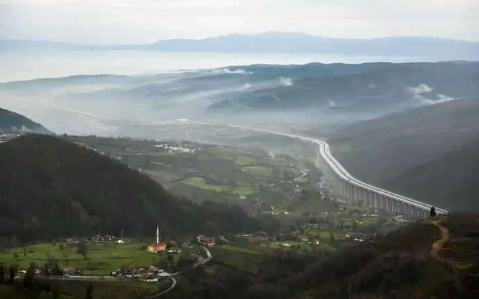 A panoramic view of Bolu, Turkey, featuring lush green valleys, scattered houses, a mosque with a tall minaret, winding roads, and a long elevated highway stretching across the mountainous landscape under a misty sky.