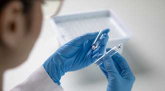 Hands wearing blue protective gloves each hold a pharmaceutical vial.