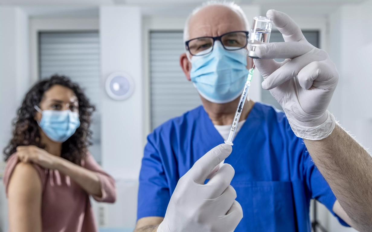 Im Vordergrund steht eine medizinisch gekleidete Person in blauer Arbeitskleidung mit Gesichtsmaske und Einweghandschuhen. Sie hält in einer Hand eine Glasampulle mit Flüssigkeit und in der anderen Hand eine Spritze. Die zweite Person hat einen Arm freigemacht, sodass der Oberarm frei für die Impfung zugänglich ist.