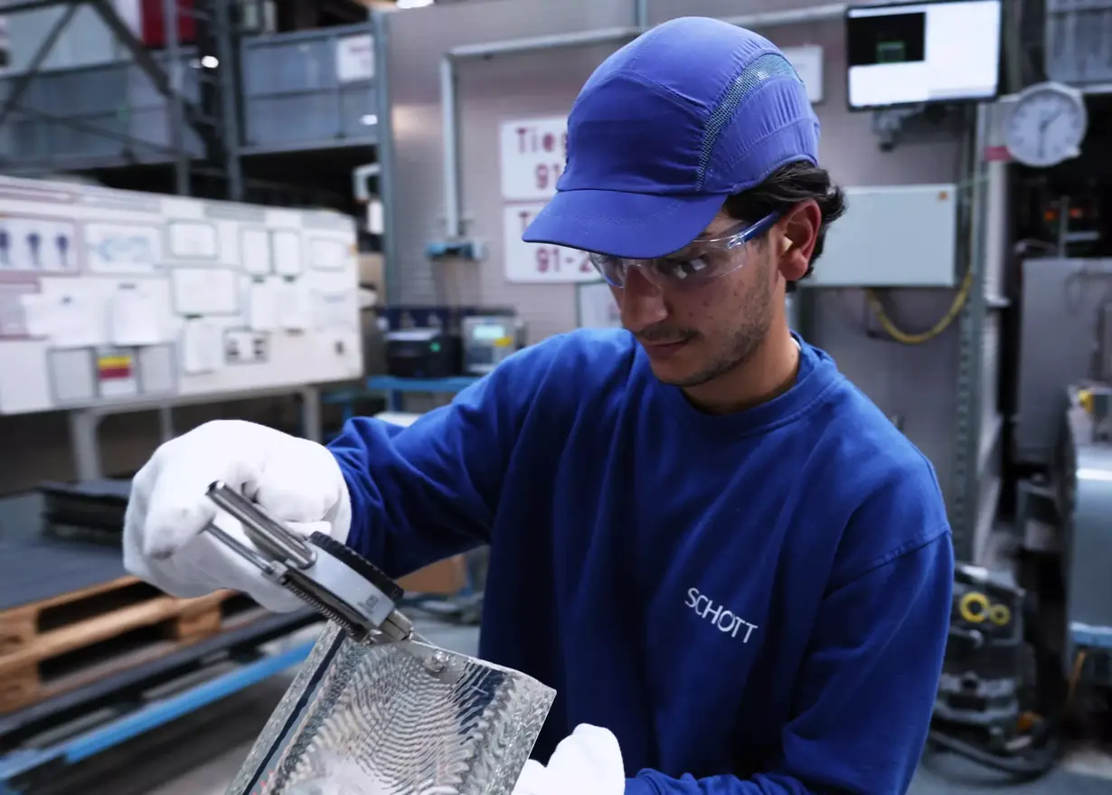 Mann in blauer SCHOTT Arbeitskleidung mit Mütze und Handschuhen bei der Kontrolle eines Glasbarrens in der Produktion.