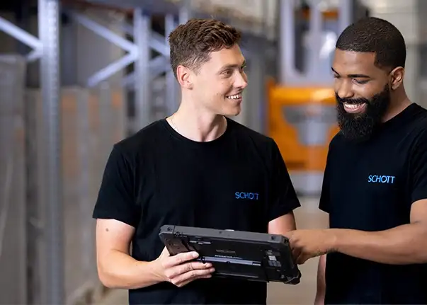 Zwei Personen in schwarzen SCHOTT‑Shirts stehen in einem Lagerbereich und betrachten gemeinsam ein Tablet.