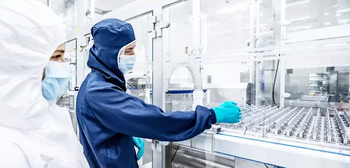 Operators in cleanroom garments monitoring and handling pharmaceutical containers on an automated production line in a sterile manufacturing environment.