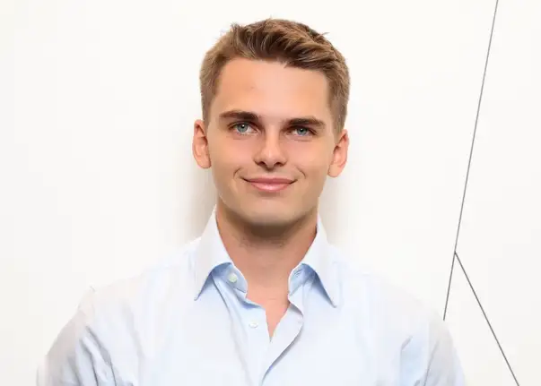 Das Bild zeigt eine Person mit kurzem Haar, die vor einer weißen Wand mit feinen Linien steht und ein helles, leicht geöffnetes Hemd trägt.