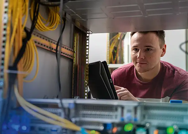 Eine Person betrachtet oder überprüft technische Geräte in einem Serverschrank und hält ein Tablet in der Hand.