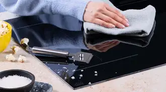 A person cleans a SCHOTT CERAN® Miradur cooktop with a cloth while salt, popcorn and kitchen utensils are placed on the countertop. 
