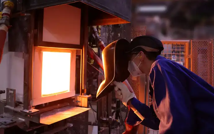 Man in heat-protective clothing and shield looking into the molten glass