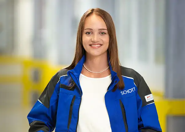 Eine Person trägt eine blaue SCHOTT‑Arbeitsjacke und steht in einer hellen Produktionshalle.
