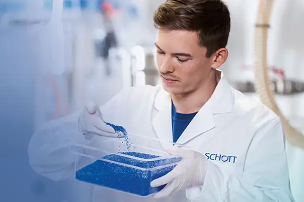Eine Person in weißem Laborkittel und Handschuhen schüttet blaue Granulatkörner in einen transparenten Behälter.