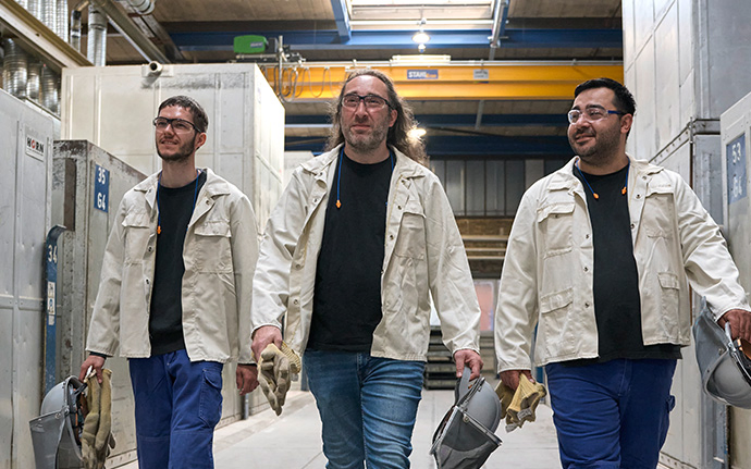 Drei Mitarbeitende in heller Schutzkleidung gehen durch eine industrielle Produktionshalle und tragen Ausrüstung in den Händen.