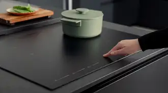 Close-up of a black matt SCHOTT CERAN® induction cooking zone with a green pot, while a hand operates the touch control panel.