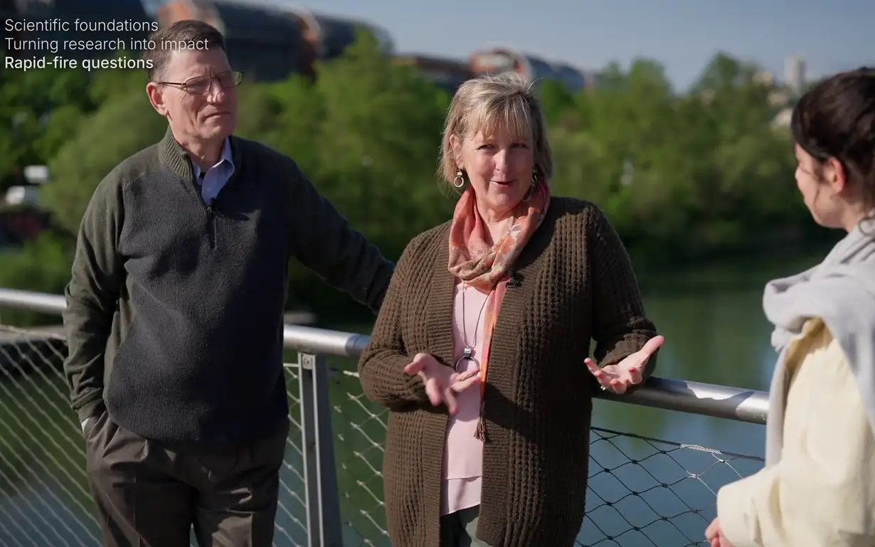 Three individuals standing on a bridge with trees and a river in the background, engaging in conversation under clear skies.
