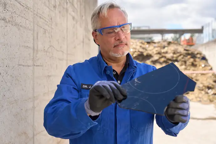 Mann in blauer SCHOTT-Schutzkleidung im Scherbenlager hält eine Glaskeramikscherbe in der Hand.