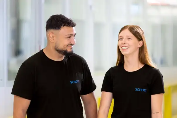 Zwei Personen in schwarzen SCHOTT‑Shirts gehen gemeinsam durch einen hellen Flur und unterhalten sich.