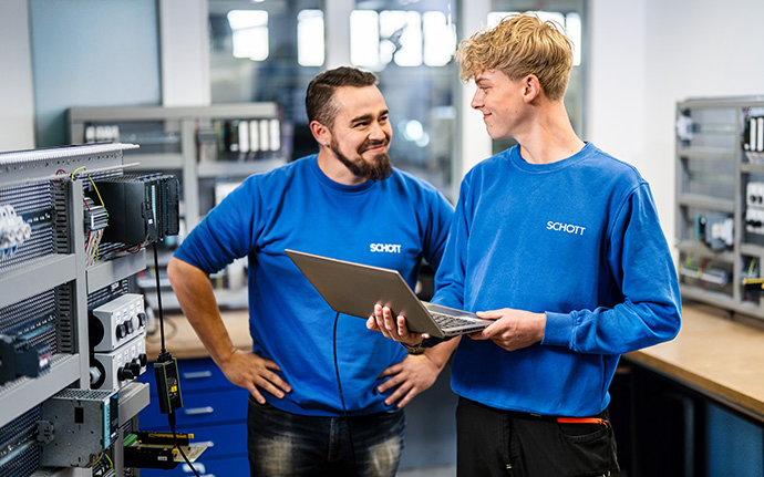  Zwei Personen in blauen SCHOTT‑Pullovern arbeiten in einer technischen Werkstatt, während eine von ihnen Daten auf einem Laptop überprüft.