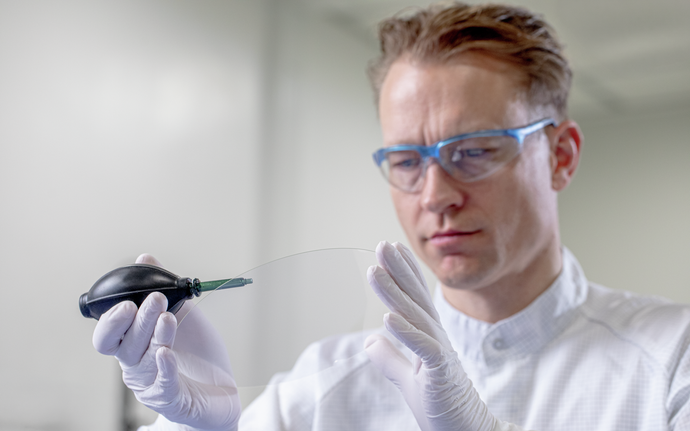 A scientist wearing protective glasses and gloves handling a thin sheet with a precision tool in a laboratory.