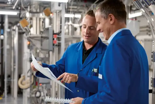 Zwei Personen in blauer Arbeitskleidung stehen in einer technischen Produktionsumgebung und besprechen gemeinsam ein Dokument mit technischen Zeichnungen.