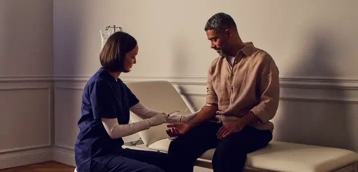 Healthcare worker drawing blood from a seated man's arm in a clinic exam room