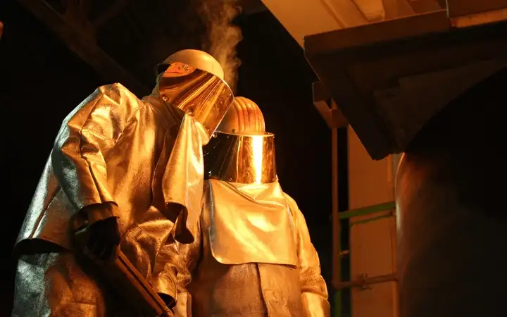 Workers in heat-resistant protective gear looking into a glass furnace.