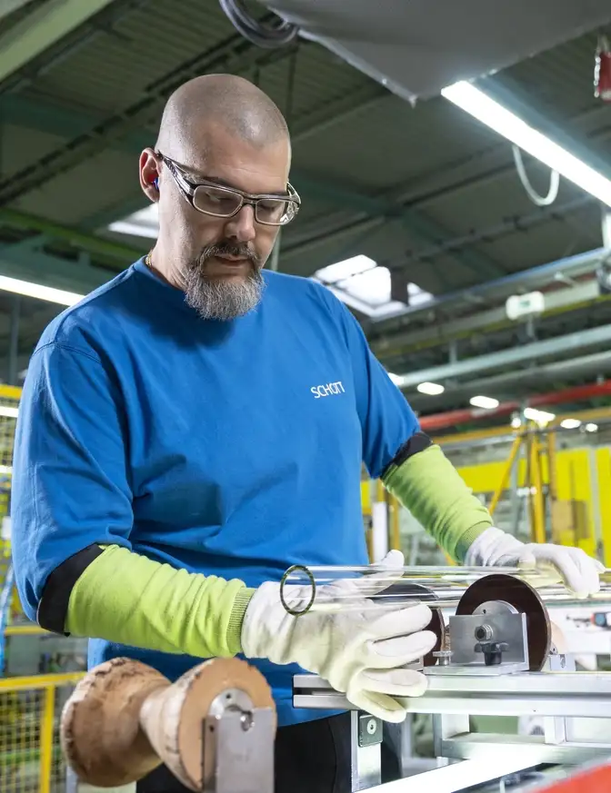Eine Person in blauer Arbeitskleidung arbeitet an einer Produktionsanlage und richtet Glasröhren aus.