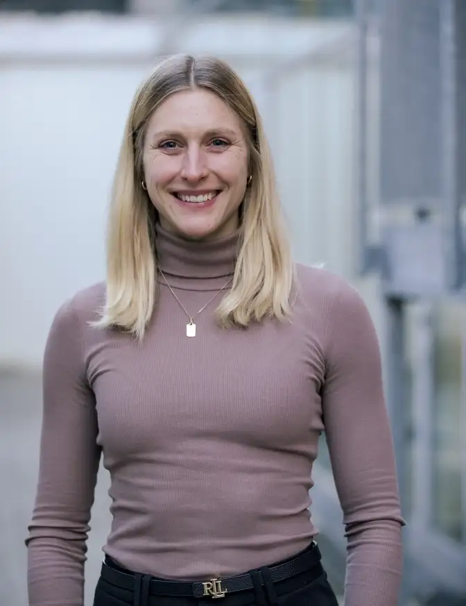 Das Bild zeigt eine Person, die im Freien vor einem hellen, unscharfen Hintergrund steht und ein langärmliges, eng anliegendes Oberteil sowie eine dunkle Hose mit einem sichtbaren Gürtel trägt, ergänzt durch eine Halsketten.