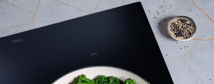 Black matte SCHOTT CERAN® cooktop surface on a light stone countertop, with a small bowl of black and white sesame seeds beside it.