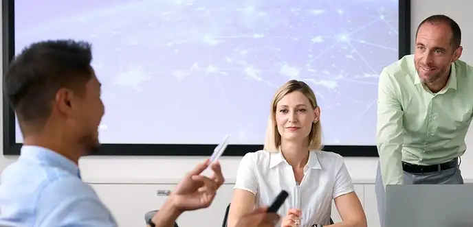 Drei Personen sitzen in einem Besprechungsraum vor einer Präsentationsfläche und diskutieren über Unterlagen und einen Laptop.