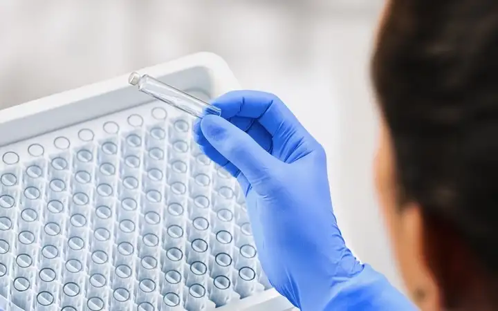 A laboratory worker in blue gloves holding a small glass vial, with a tray of vials in the background.