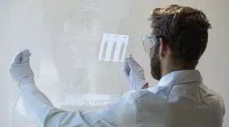 Man in cleanroom clothing holding a glass panel in his hands.