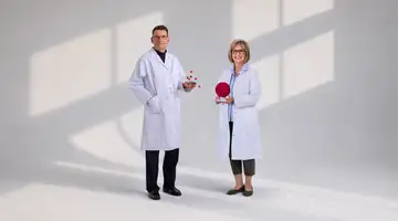 Two individuals wearing lab coats stand in a well-lit space holding scientific models, with a shadow of window panes in the background.