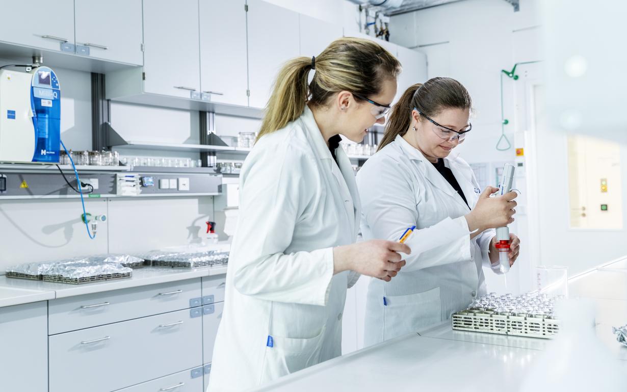 Zwei Frauen im Labor mit Schutzbrille und weißen Arbeitskitteln bei der Prüfung eines Pharmafläschchen mittelns SCHOTT Pharma’s PartnerLab.