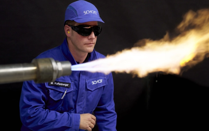 Man in blue SCHOTT work clothes with safety goggles behind a flame.