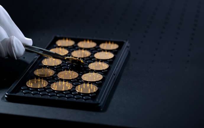 Gloved hand with tweezers handling black plate with gold-plated sensor feedthroughs.