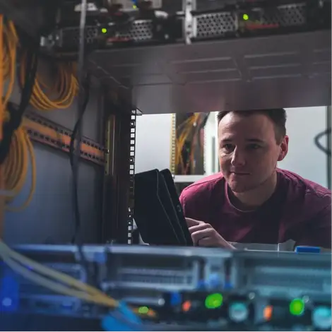 Eine Person betrachtet oder überprüft technische Geräte in einem Serverschrank und hält ein Tablet in der Hand.