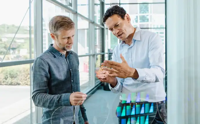 Two men examine a transparent object behind a counter with optical displays, set in a bright showroom with large windows.