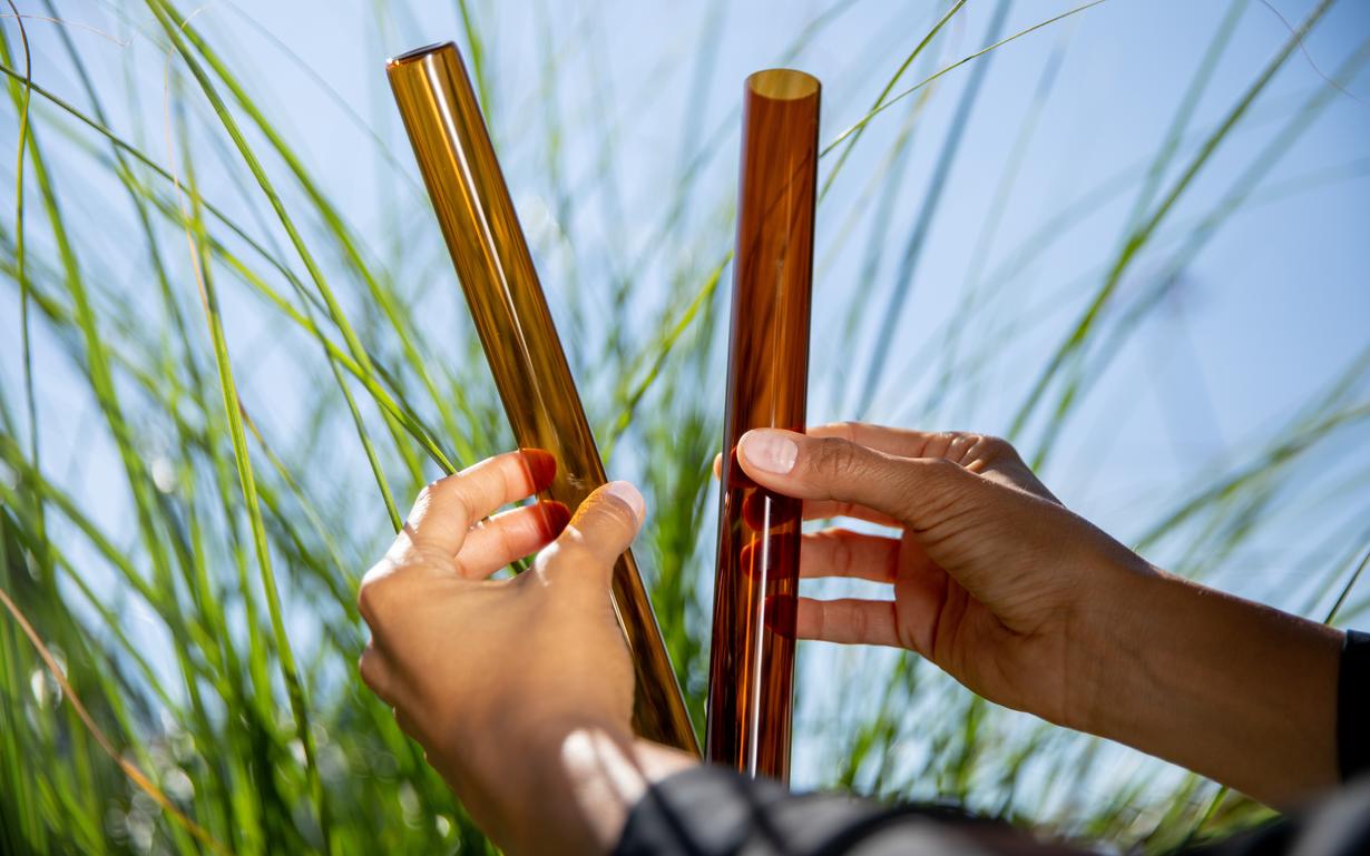 Two hands holding two brown glass tubes, against blue sky and grass background.