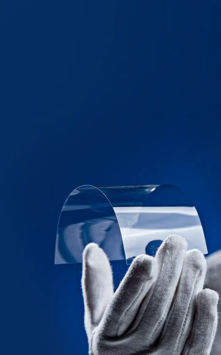 A hand in white gloves holding a thin, flexible transparent sheet against a blue background.