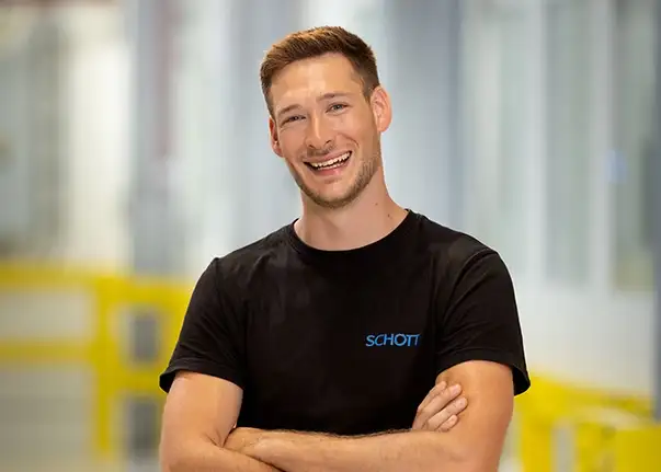 Eine Person trägt ein schwarzes SCHOTT‑T-Shirt und steht mit verschränkten Armen in einer hellen Produktionsumgebung.
