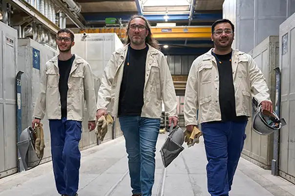 Drei Personen in Arbeitskleidung gehen nebeneinander durch eine industrielle Produktionshalle und tragen Schutzausrüstung in den Händen.