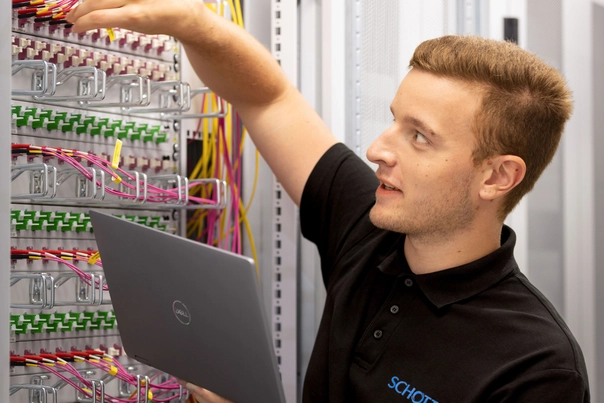 Mann mit Laptop in Serverraum steckt Stecker in eine große Maschine.