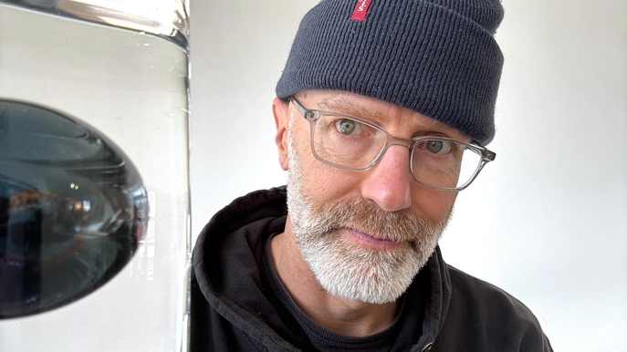 Man with glasses, white beard, and navy beanie posing beside a tall glass container indoors