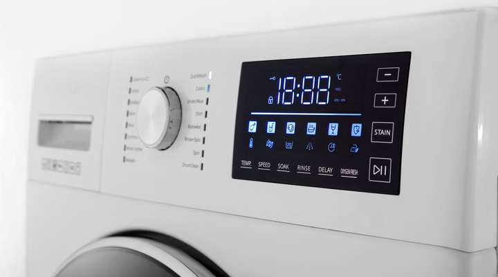 A close-up view of a modern washing machine's control panel made of glass, central dial with labeled wash options like "QuickWash" and "Cottons", and buttons.